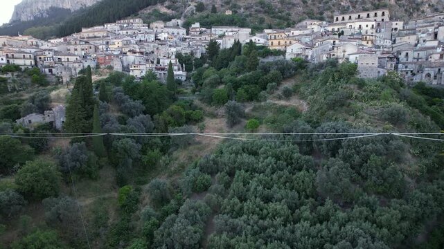 Drone footage shows a highline sling strung over a mountain valley. The highline sling and rigging are visible strung over the valley against the backdrop of a picturesque Italian hillside town.