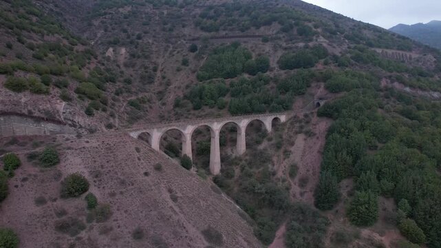 Stone viaduct bridge spans green mountain valley with forested hills and rocky slopes under cloudy sky for travel infrastructure landscape adventure nature exploration drone photography footage 