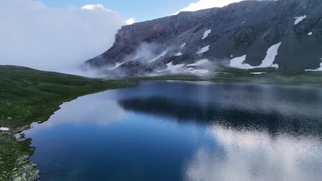 Cinematic aerial view of a misty alpine lake with low clouds rolling over mountain cliffs and snow patches