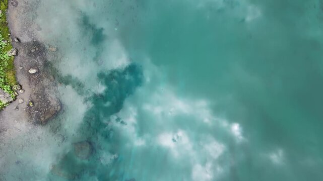 Top-down aerial view of a crystal clear turquoise mountain lake water with reflections of clouds
