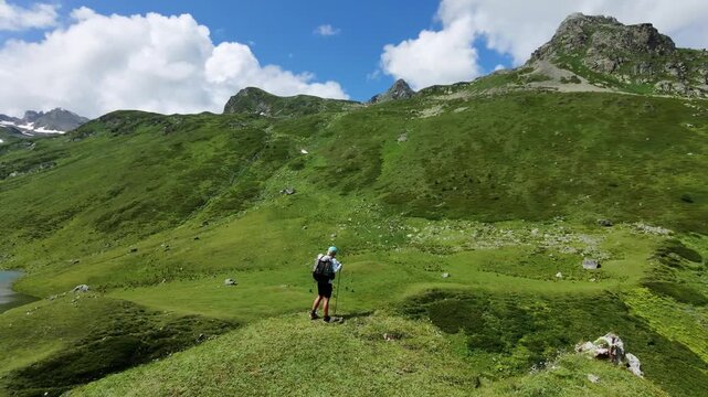 Drone shot of a tourist trekking on a green alpine slope, standing on a mountain summit in summer