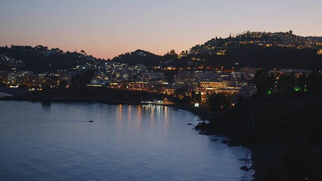 Night time lapse of Almunecar bay and city lights with calm Mediterranean sea and small boat approaching shore. 