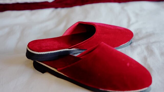 Pair of red velvet groom slippers with black soles placed neatly on white bed sheet for traditional wedding ceremony angled close up shot