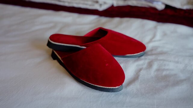 Wide shot of red velvet wedding slippers resting on crumpled white bedsheet with soft natural light and blurred decorative fabric in background