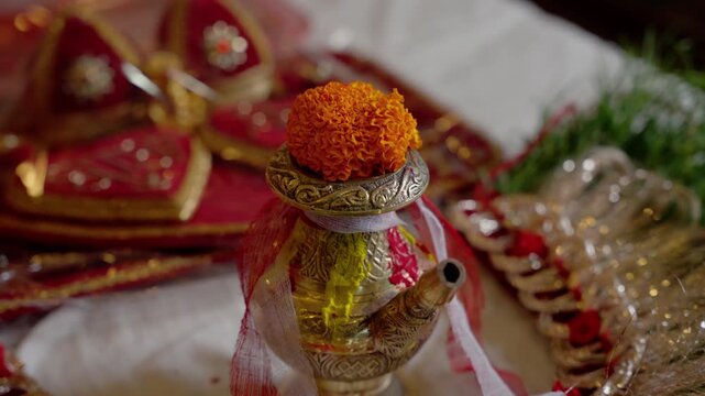 Close up of ornate brass kalash sacred pot topped with orange marigold flower and turmeric tika during Hindu wedding ritual preparation indoors