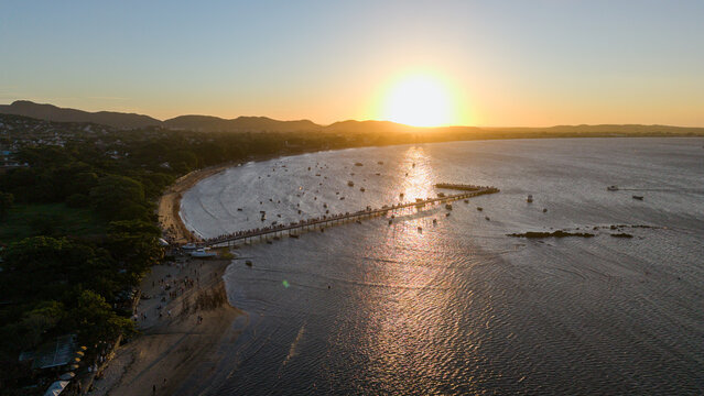 Aerial view of Praia de Manguinhos at sunset with a long pier, anchored boats, and people on the sandy shore, Armacao dos Buzios, Rio de Janeiro, Brazil.