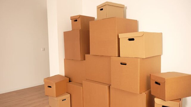 Cardboard boxes stack in corner. Sizes vary from small to large. White walls and wooden floor surround. Stacks suggest moving or storage. Ideal for home organization scenes