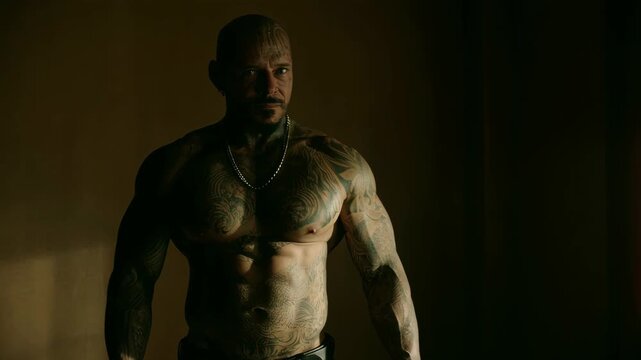 Muscular tattooed man standing in dimly lit room with intense expression