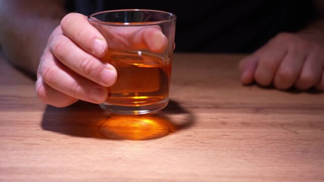 Hand grips a small glass with amber liquid. Wooden table shows natural texture. Person's expression is calm and steady. Ideal for health or beverage promotion content