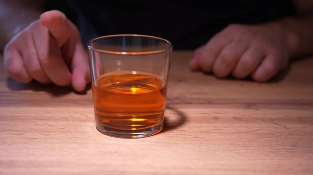 An alcoholic at a table in front of a glass of whiskey tries to resist the urge to drink a glass of alcohol. Alcoholism.