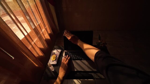 POV of a person cleaning a laptop screen with microfiber cloth and spray cleaner in a warm morning light room, slow motion 4K 120fps.