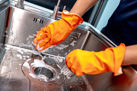 Housekeeper hand wearing orange rubber glove scrubbing on kitchen sink surface and drainer with soap suds spreading around. Cleaning stainless steel sink for hygiene. Housework chores in the kitchen