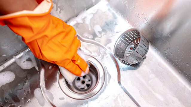 Housekeeper hand wearing orange rubber glove scrubbing on kitchen sink drainer with soap suds spreading around. Cleaning stainless steel sink for hygiene. Housework chores in the kitchen