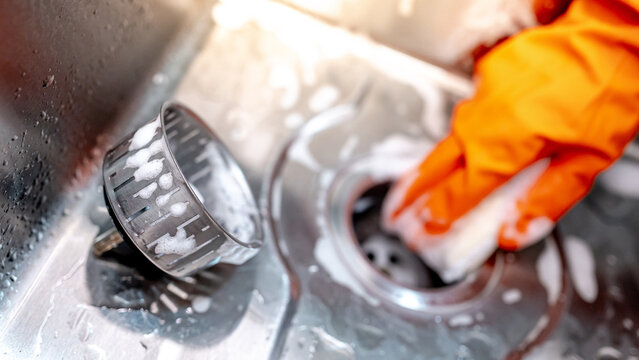 Housekeeper hand wearing orange rubber glove scrubbing on kitchen sink drainer with soap suds spreading around. Cleaning stainless steel sink for hygiene. Housework chores in the kitchen