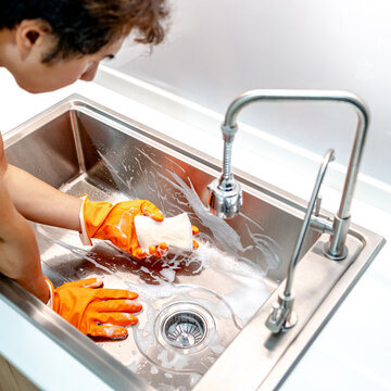 Asian male housekeeper with orange rubber glove scrubbing on kitchen sink surface with soap suds spreading around. Cleaning stainless steel sink for hygiene. Housework chores in the kitchen