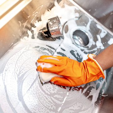 Housekeeper hand wearing orange rubber glove scrubbing on kitchen sink drainer with soap suds spreading around. Cleaning stainless steel sink for hygiene. Housework chores in the kitchen