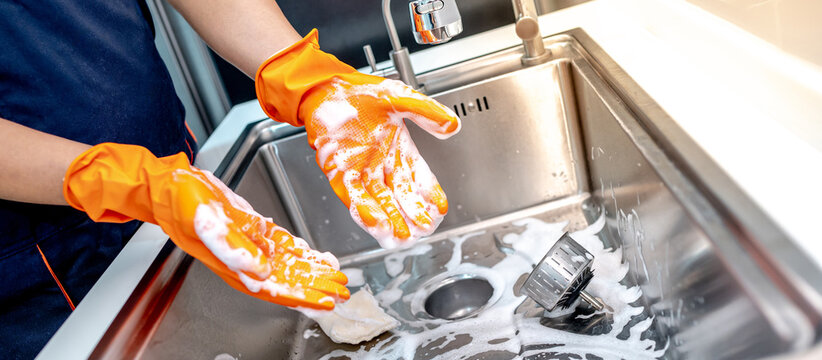 Housekeeper showing hands with orange rubber glove full with soap suds while scrubbing on kitchen sink surface. Cleaning stainless steel sink for hygiene. Housework chores in the kitchen