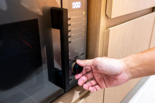 Hand turning knob setting timer on modern black microwave oven in wooden cabinet. Using home appliance in the kitchen