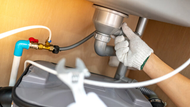 Plumber hand with glove fixing pipe connection with adjustable wrench and checking grease trap box under kitchen sink. Handyman repairing plumbing system in wooden cabinet.