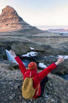 Portrait of a woman sitting with her arms raised by the Kirkjufell mountain waterfall, Iceland.