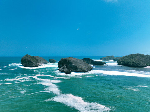 Aerial view of Pantai Kasap with rocky islands, turquoise sea, white waves, and clear blue sky in Kecamatan Pringkuku, Jawa Timur, Indonesia.