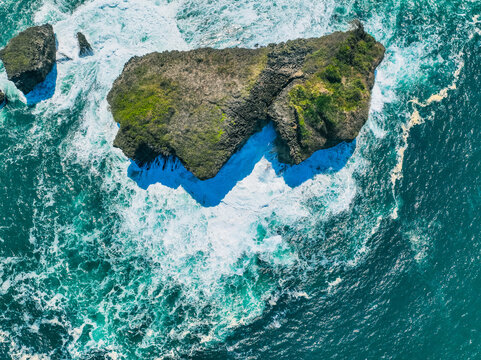 Aerial view of Pantai Kasap with its rocky cliffs and turquoise ocean waves crashing against the shore in Kecamatan Pringkuku, Jawa Timur, Indonesia.