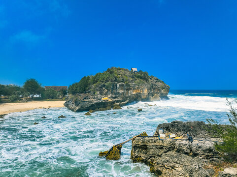Aerial view of the rocky islet at Pantai Kasap surrounded by crashing waves, sandy beach, and lush vegetation under a clear blue sky in Kecamatan Pringkuku, Jawa Timur, Indonesia.
