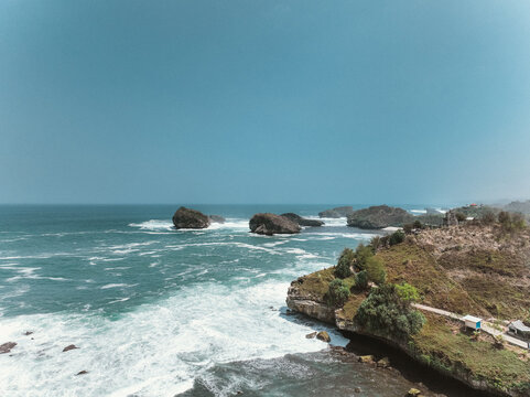 Aerial view of Pantai Kasap featuring dramatic rocky cliffs, turquoise ocean waves crashing against the shore, and lush coastal vegetation in Kecamatan Pringkuku, Jawa Timur, Indonesia.