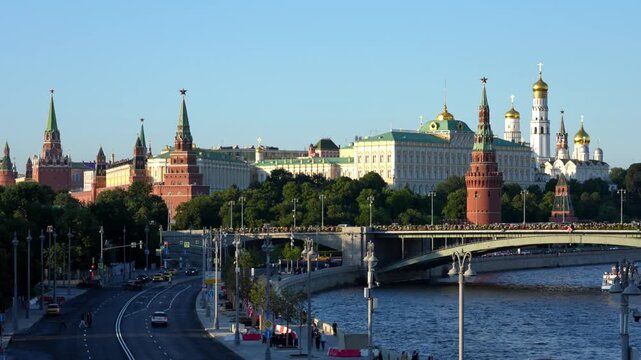 Moscow, August 10, 2026, Clear blue sky highlights Moscow's Kremlin and St. Basil's Cathedral. The Moskva River flows beneath a crowded bridge. Pedestrians smile while taking photos of the city