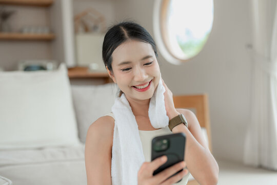 Asian woman in white activewear with a towel around her neck smiles at her phone after exercising in a bright living room.