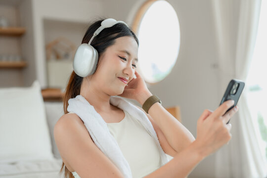 Asian woman in beige activewear wearing headphones and a towel smiles at her phone after a home workout in a bright living room.