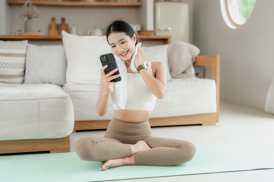 Asian woman in beige activewear with a towel around her neck sits on a yoga mat and smiles at her phone after a home workout.