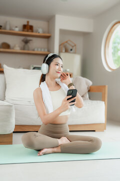Asian woman in beige activewear wearing headphones and a towel sits on a yoga mat and listens to music on her phone with a smile.