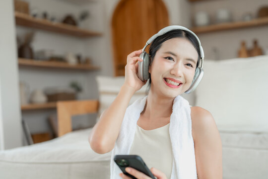 Asian woman in beige activewear wearing headphones and a towel smiles at the camera while holding her phone after a home workout.