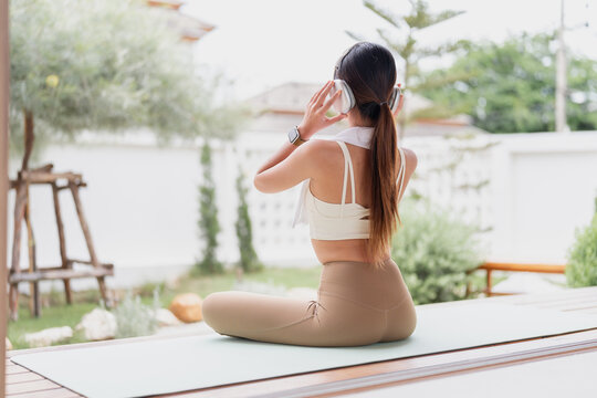 Back view of an Asian woman in activewear putting on headphones while sitting on a yoga mat on a terrace with garden views.