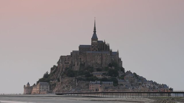 Mont Saint Michel abbey rising above the tidal flats at dusk, Normandy, France. Static shot of the medieval island town, spire and ramparts under a soft pink sky at low tide in summer