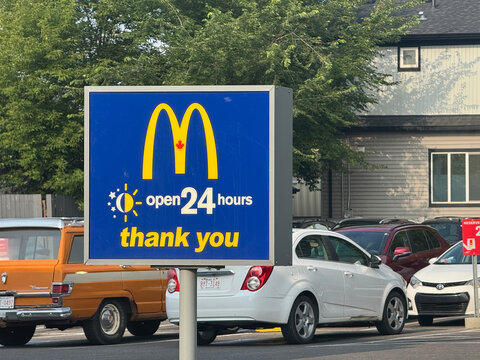 Calgary, AB, Canada. August 16, 2026. Medium view of a blue McDonald's open 24 hours sign in front of a parking lot filled with parked cars.