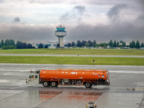 Ottawa, ON, Canada. August 15, 2026. Bright orange fuel tanker truck parked on tarmac near airfield control tower under cloudy skies.