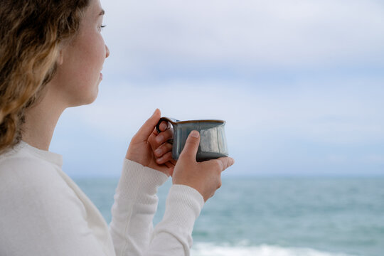 A woman enjoying a warm drink while gazing at the ocean on a cloudy day. A quiet seaside moment, a slow life and mindful relaxation in cool weather.