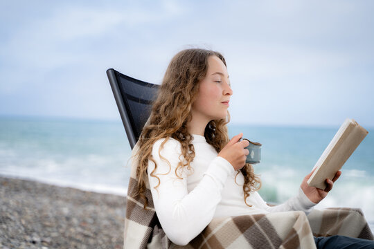 Woman enjoying a quiet seaside moment with a book and hot beverage outdoors. Cozy off-season travel, slow living and mindful relaxation by the ocean.