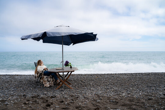 Wide coastal lifestyle scene with a woman resting under a beach umbrella beside the sea. Quiet off-season vacation and seaside relaxation concept.