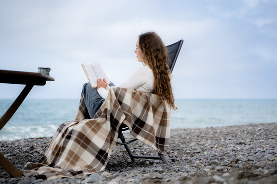 Woman immersed in a printed book while resting on a pebble beach by the ocean. Slow living, reading and peaceful seaside retreat concept.