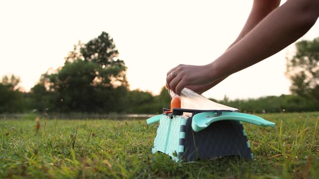 Portable folding plastic toilet for child stands beside river at sunset. Slow motion camera approaches while female hands raise water filled bag above potty.
