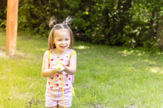 Young girl with pigtails wearing a pink heart-patterned tank top and shorts plays with a water toy in a grassy backyard during a sunny day