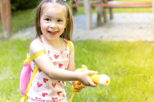 Young girl with long hair wearing a pink heart-patterned shirt holds a yellow water gun while playing outdoors in a grassy area with playground equipment visible