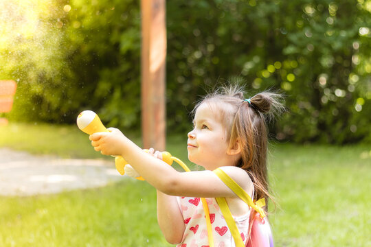 Young girl with brown hair in pigtails sprays water from a yellow toy water gun while playing in a green park on a sunny day with trees in the background