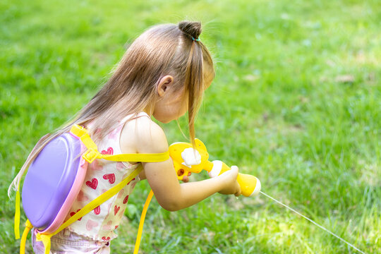 Young girl with long hair wearing a pink and white outfit uses a yellow water gun while playing on green grass in a sunny outdoor setting
