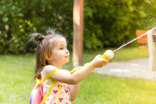 Young girl with brown hair in pigtails sprays water from a yellow garden hose while playing in a green park with trees and a playground visible in the background