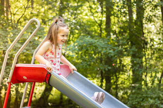Young girl with brown hair wearing a pink dress slides down a playground slide surrounded by green trees in a sunny outdoor park setting