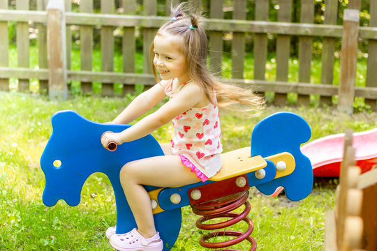Young girl with long hair wearing a white tank top with hearts rides a blue spring horse in a sunny playground surrounded by a wooden fence and green grass